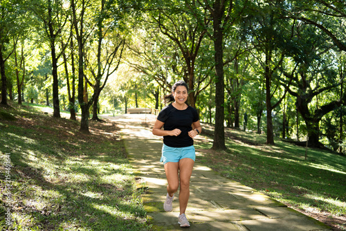 Woman, running and smiling in park with fitness, wide shot or trail path outdoor. Front view, athlete or runner with smart watch, nature and cardio training in forest