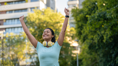 Woman, celebrating and arms up in city with fitness, achievement or high energy outdoor. Athlete or runner with headphones, smart watch and enthusiasm for workout success