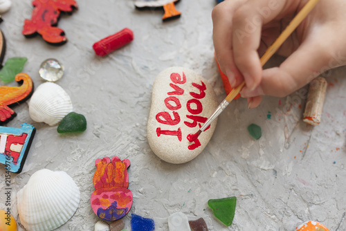 Child hand painting small red heart next to I love you text on stone