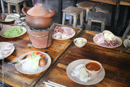 Traditional Thai Jim Jum hot pot served in a clay pot with fresh vegetables, pork, tofu, and dipping sauce on a wooden table.