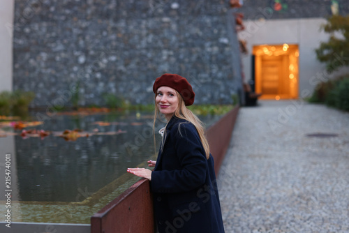 Fashionable young woman wearing red beret and black coat standing near water in an urban setting during autumn.