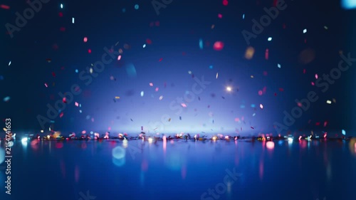 Small bits of colorful paper fall slowly through a blue space. The reflective floor creates a double image of the falling confetti. 3D render motion.