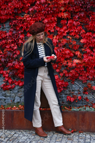 Stylish young woman in beret and long coat holding a red autumn leaf against vibrant red foliage.