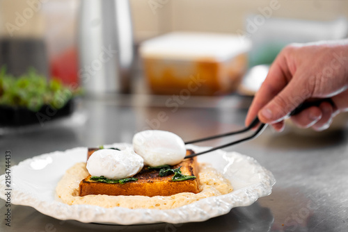 Obraz na płótnie A hand using tweezers to place a poached egg on a piece of toasted brioche bread, with greens on top, served on a white plate over a creamy sauce in a professional kitchen