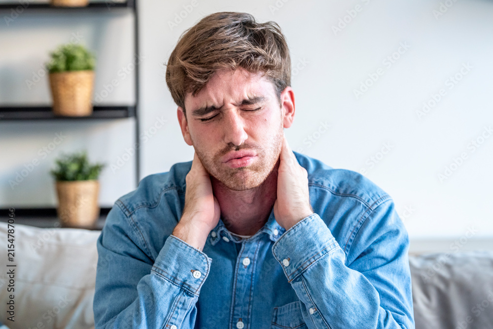 Fototapeta premium Young man with neck pain holding his neck, wincing in discomfort on a couch at home