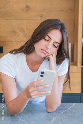 Canvas Print Young woman in white t-shirt looking bored and scrolling smartphone at kitchen c