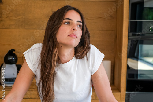 Foto Young woman in white t-shirt leaning on kitchen counter looking thoughtful and p