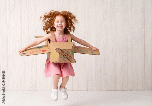 Cute redheaded child playing pilot with cardboard plane against white wooden background