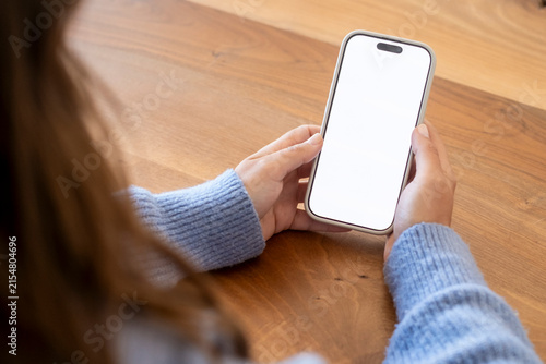 Person holding smartphone with blank white screen over wooden table, hands in blue sweater ready to use mobile phone