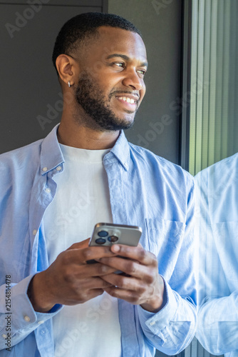 Quadro su tela Young man smiling and holding smartphone while looking out a window, wearing a c