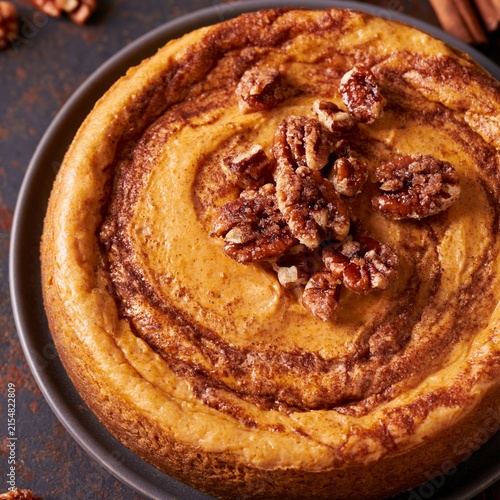 Pumpkin cheesecake topped with caramelized pecans on a dark plate, surrounded by scattered nuts and cinnamon sticks, overhead view showcasing dessert details