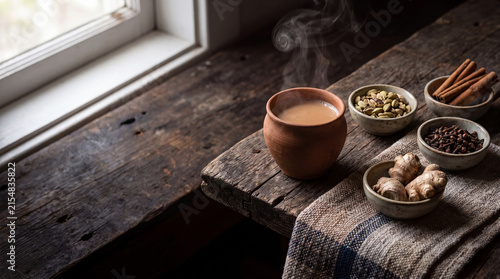 Steaming cup of indian chai tea with spices including cinnamon sticks, cardamom pods, cloves, and ginger on rustic wooden surface keywords: chai tea, hot drink, beverage