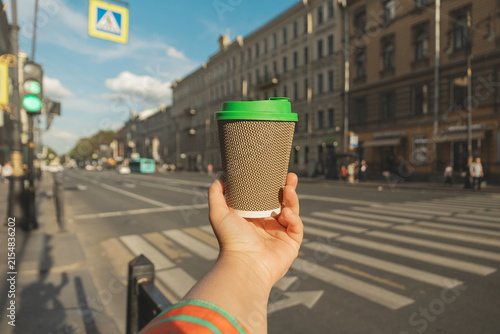 Hand holding ripple coffee cup with green lid on blurred city street background