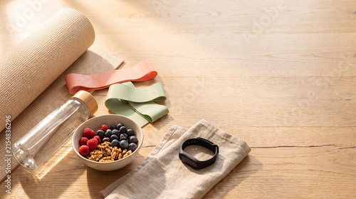 Yoga mat water bottle resistance bands and a bowl of berries and nuts on a wooden surface with soft light