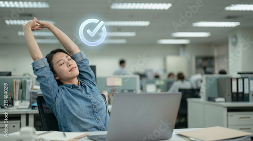 Young woman stretching arms up at desk in office, taking a break, feeling refreshed with a digital checkmark overlay indicating task completion, blurred colleagues in background, sense of