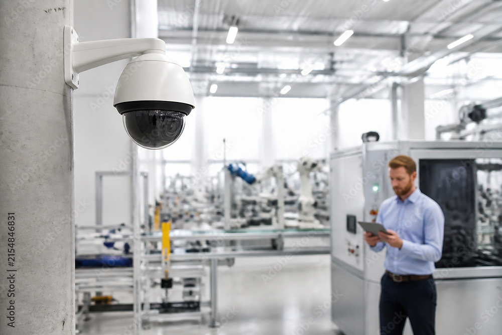 Fototapeta premium A security camera is mounted on a pillar in a bright, modern factory setting where a man is looking at a tablet.