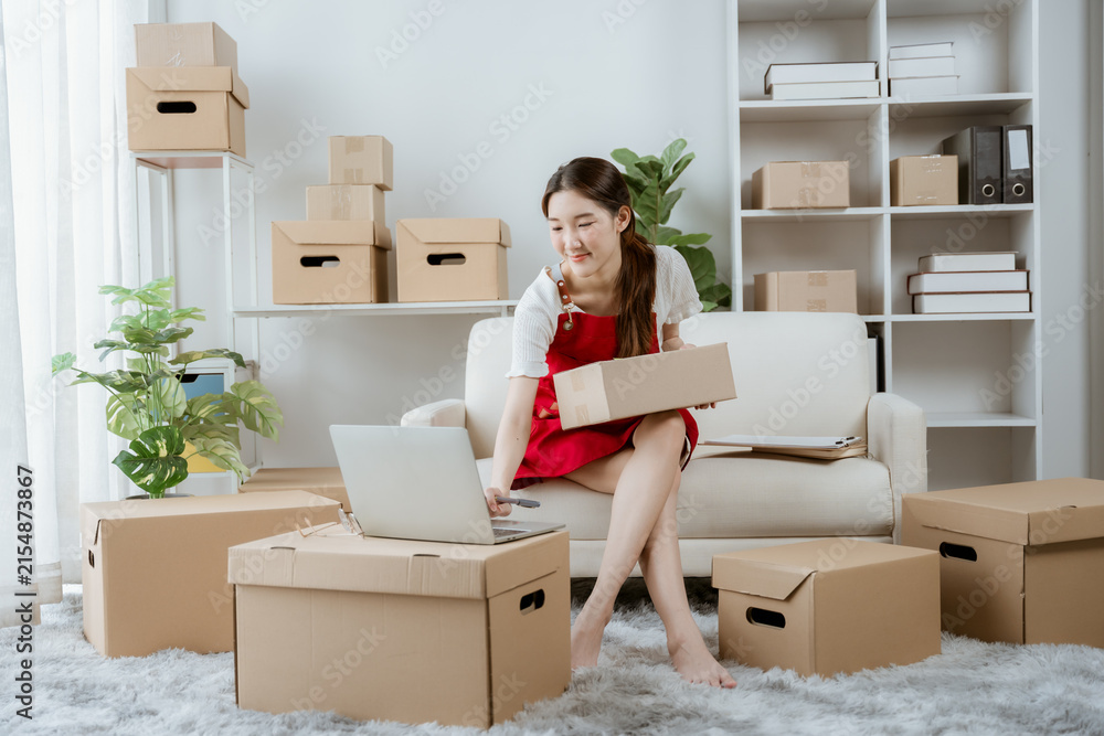 Fototapeta premium Woman Preparing Packages for Shipping: A young woman sits in a well-lit room surrounded by cardboard boxes, laptop open on her lap, as she meticulously prepares items for shipment.