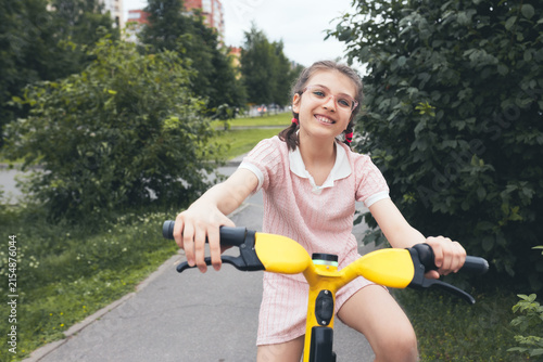 Happy young girl in glasses riding yellow electric scooter in summer park