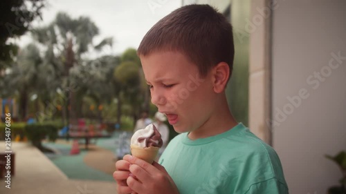 Young boy enjoying a chocolate ice cream cone outdoors, happily licking the melting treat. Sunny summer setting with a pool and greenery in the background, capturing a joyful childhood moment.