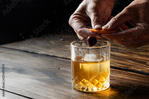 Hands pressing an orange peel releasing the oils on top of an old fashioned cocktail on a wooden bar top.