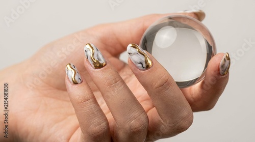 High-resolution close-up photo of a woman's manicured hand holding a clear transparent glass sphere crystal ball, displaying luxurious white marble fingernails decorated with 3D metallic liquid gold c