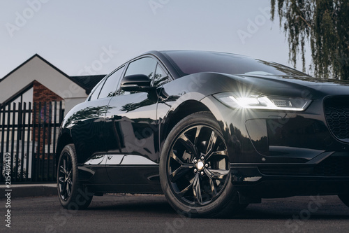 A glossy black car with illuminated headlights is parked on a paved driveway, reflecting nearby trees and buildings in its polished bodywork.