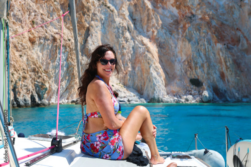 A smiling woman wearing sunglasses and a floral swimsuit relaxes on a sailboat near Milos, surrounded by calm blue water and bright Mediterranean sunlight.