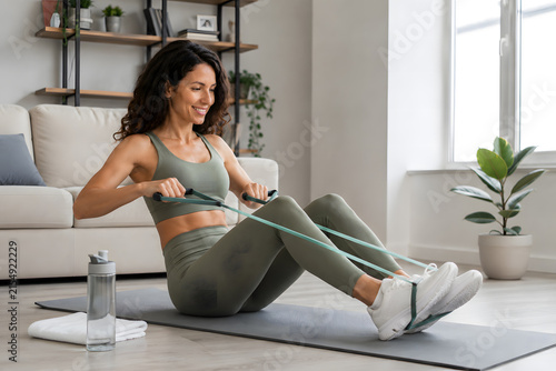Plano entero de mujer sonriente entrenando en casa con banda elástica sobre esterilla de yoga con luz natural en ángulo medio.