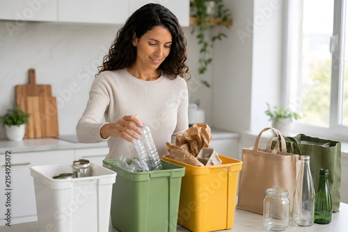 Plano medio de mujer sonriente clasificando botellas de plástico y papel en contenedores de colores en cocina brillante con luz natural.