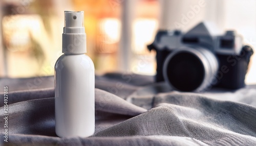white plastic cosmetic spray bottle standing on a soft textured fabric cloth with a blurred camera in the bright background