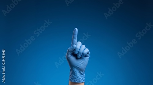 A hand wearing a blue medical glove raises a single index finger against a plain blue background, signifying 'one' or 'number one'.