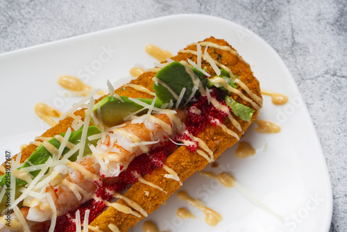 High angle view of crispy sushi hot dog roll stuffed with whole tiger shrimp, avocado and red tobiko caviar. Deep fried crunch sushi roll with shrimp on a white plate against concrete background, copy