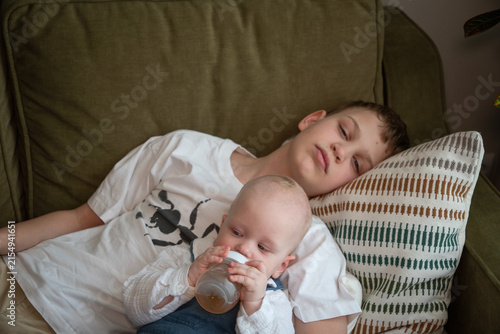 Obraz na płótnie Older brother watches over baby sibling while younger child drinks from bottle resting in his arms on sofa at home