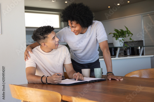 Diverse male friends sitting at kitchen table, leaning over and reviewing binder with mugs