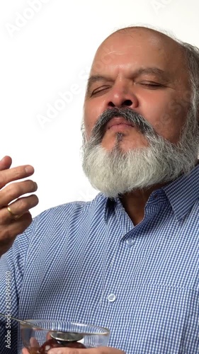 An Indian Asian elderly businessman with a beard is happily enjoying gulabjamun from a glass bowl with eyes closed , expressing great pleasure with hand gestures 