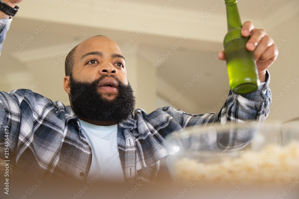 Fototapeta premium African American man in flannel shirt sitting home holding green bottle near popcorn bowl reacting