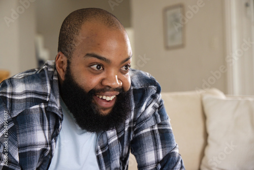 Fotografie African man leaning forward, smiling wide-eyed on beige couch at home with plaid