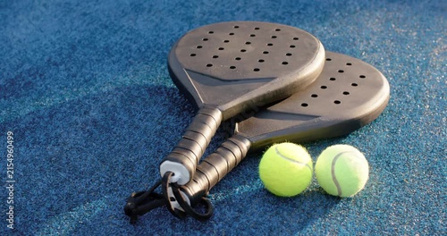 Camera and sunlight are shifting to reveal stacked black paddles and yellow balls on blue court
