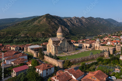 Historic Mtskheta cityscape with Svetitskhoveli Cathedral, traditional Georgian houses, and forested mountains under a clear blue sky
