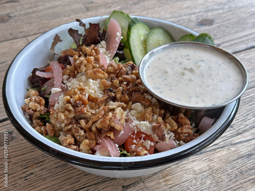 Konstfotografi A mixed greens salad with grape tomatoes, cucumber, pickled red onions, honey walnuts, feta, and creamy lemon and feta dressing is shown in a bowl on a restaurant table