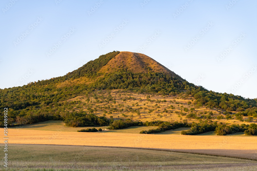 Obraz premium Oblik Hill stands tall in the Bohemian Central Mountains of Czechia. The sun shines on the green slopes and fields nearby. Visitors can enjoy nature and the surrounding landscape.