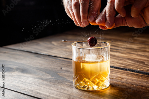 Hands pressing an orange peel releasing the oils on top of an old fashioned cocktail on a wooden bar top.