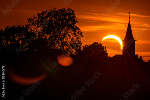 Partial solar eclipse above a church silhouette at sunset, with dramatic orange sky, dark trees and lens flare creating a mysterious atmospheric scene.