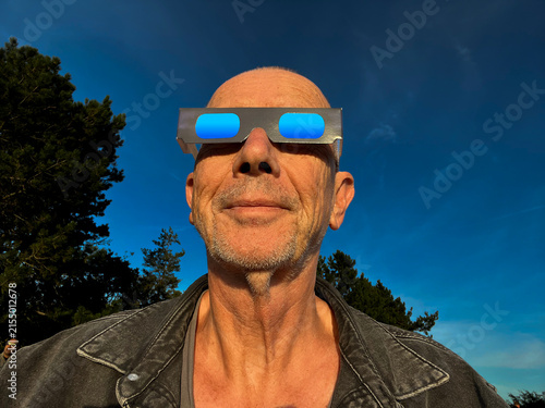 Man looking at the solar eclipse with cardboard protection eclipse glasses with blue sky background.