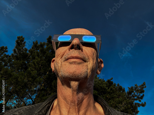 Man looking at the solar eclipse with cardboard protection eclipse glasses with blue sky background.