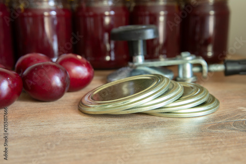 Close-up of several metal canning jar lids arranged on a wooden table, with glass jars of homemade preserves softly blurred in the background. The image illustrates the process and preparation of home