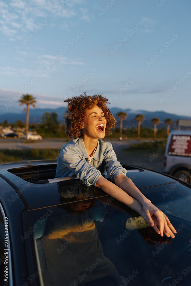 Obraz premium Candid lifestyle portrait of a joyful woman leaning from a car window, outdoors at golden hour, palm trees in the background, capturing carefree travel mood and radiant happiness