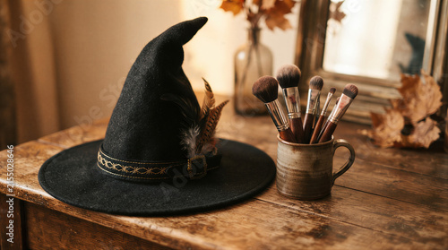 Witch hat with feather beside makeup brush in rustic interior. Halloween costume and beauty concept with autumn decoration, creative seasonal still life and copy space.