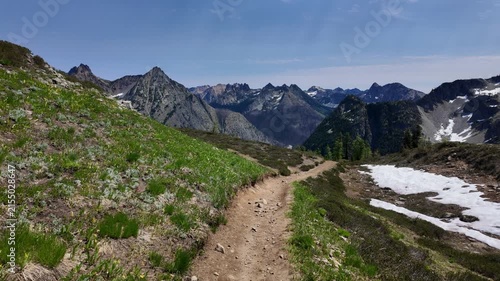 First-person point of view moving forward along a dirt mountain path with snowfield patches and dramatic mountain range in the background on a sunny day in the North Cascades, Washington