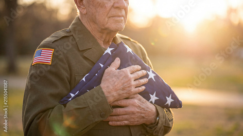 An elderly US soldier in uniform lovingly holds a folded American flag against his chest as the sun sets behind him.
